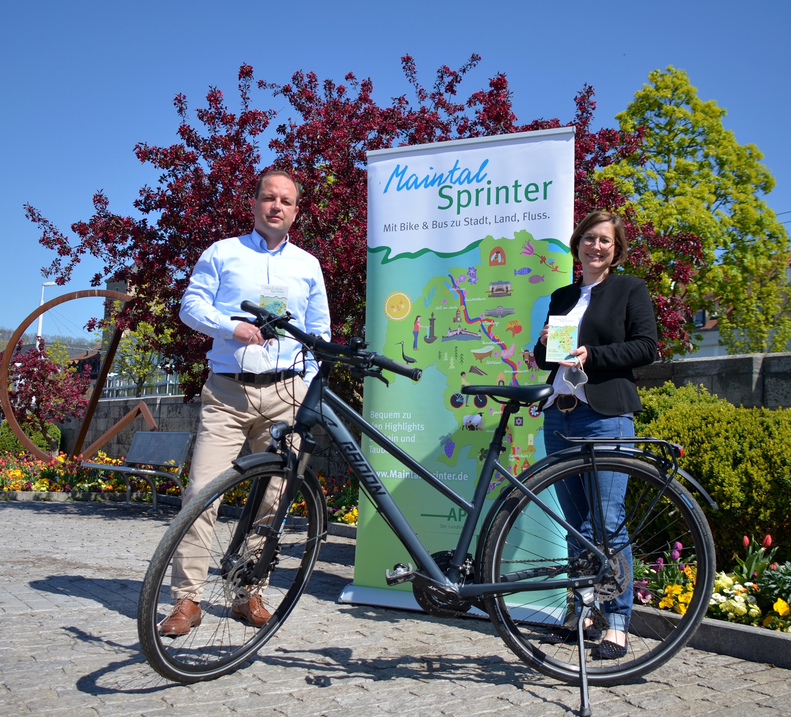APG-Betriebsleiter Dominik Stiller und Dr. Sibylle Holste, stv. APG-Betriebsleiterin stehen hinter einem Fahrrad und vor einer Streckenkarte des Maintalsprinter.